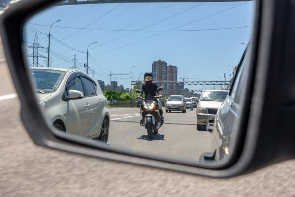 The reflection of a motorcycle in the left side mirror of a car traveling in traffic and preparing to overtake between two cars in a narrow side distance at speed.